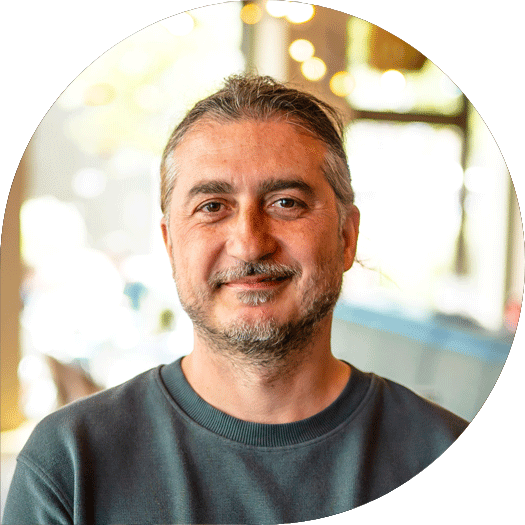 Porträt eines Mannes mit dunklem, zurückgebundenem Haar und Dreitagebart in einem dunkelgrauen Pullover, vor einem unscharfen Hintergrund mit warmem Licht.