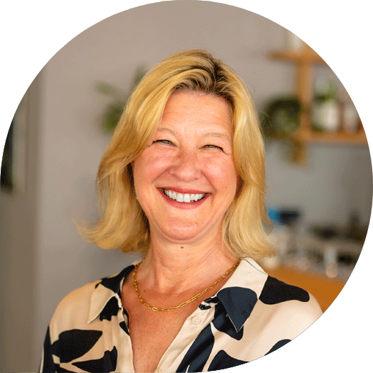 Porträt einer fröhlich lächelnden Frau mit blondem Haar und gemusterter Bluse, vor einem warm gestalteten Hintergrund mit Pflanzen und Regalen.