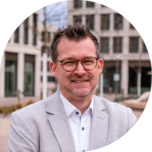 Porträt eines Mannes mit Brille und kurzem, dunklem Haar im hellgrauen Sakko, vor dem modernen Gebäudeensemble der Heidelberger Volksbank.