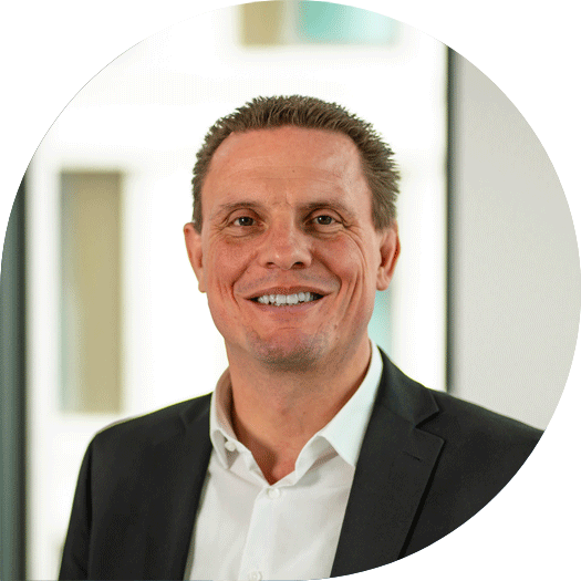 Porträt eines Mannes im dunklen Anzug und weißem Hemd, lächelnd vor einem Fenster mit moderner Gebäudefassade im Hintergrund.