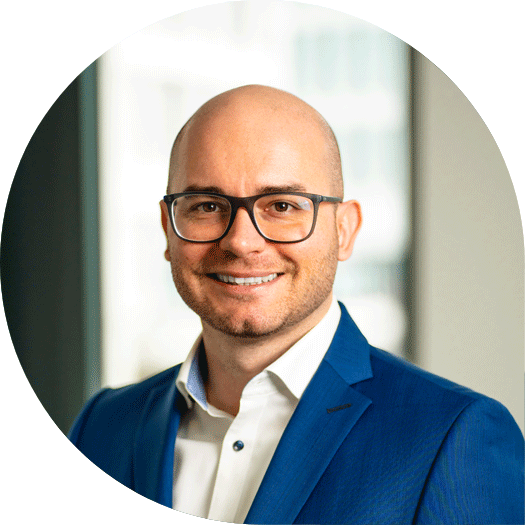 Porträt eines lächelnden Mannes mit Brille, weißem Hemd und blauem Anzug vor einem Fenster mit städtischem Hintergrund.