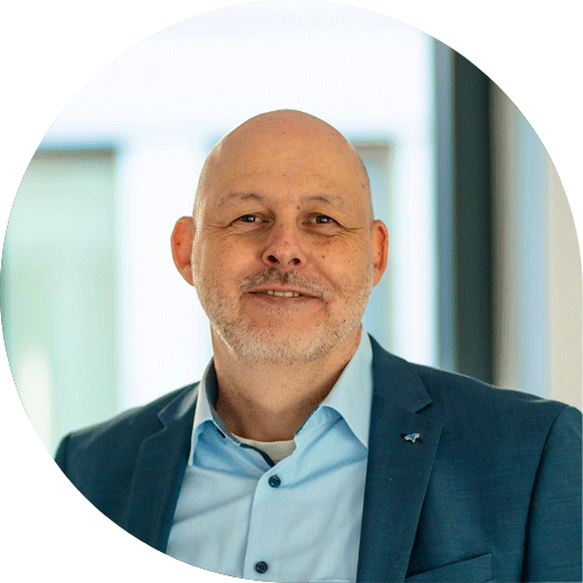 Porträt eines Mannes mit Glatze und grauem Bart, im blauen Anzug und hellblauen Hemd, vor einem lichtdurchfluteten Fensterbereich.