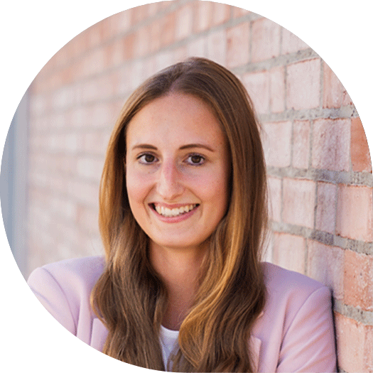 Porträt einer jungen Frau mit langen, hellbraunen Haaren, die lächelnd vor einer Backsteinwand steht und einen rosa Blazer trägt.