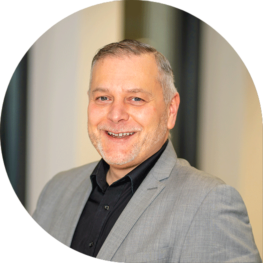 Porträt eines Mannes mit grauem Sakko und schwarzem Hemd, lächelnd vor einem hellen, modernen Hintergrund.