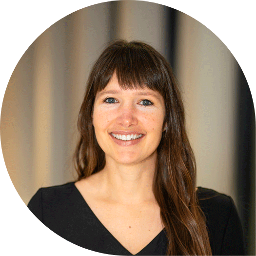 Porträt einer lächelnden Frau mit langen braunen Haaren in schwarzer Kleidung, vor neutralem Hintergrund.